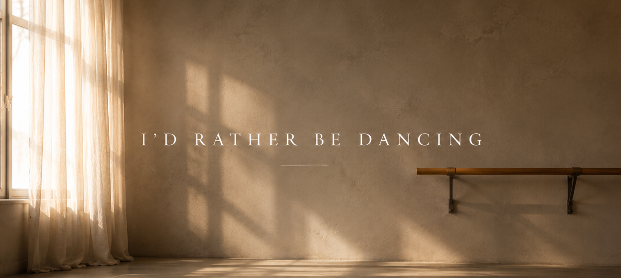 A minimalist, empty dance studio with warm sunlight streaming through a window, casting shadows on a textured beige wall and concrete floor. A wooden ballet barre is mounted on the wall, and the text 'I'D RATHER BE DANCING' is displayed in the center.