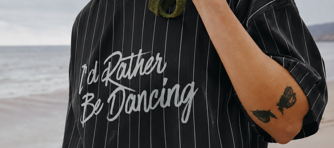 Person wearing a black pinstripe shirt with 'I'd Rather Be Dancing' text on a beach.